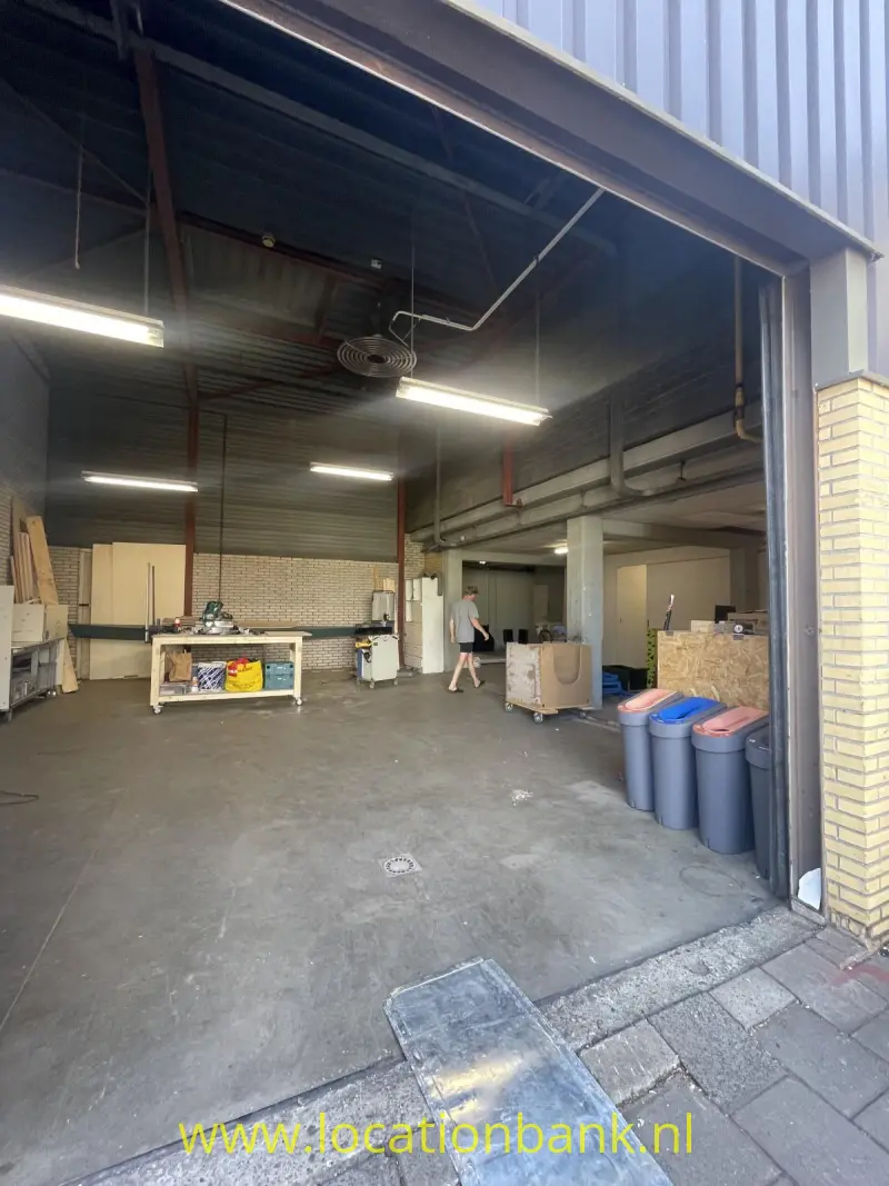 Een open hal met een metalen dakconstructie, werkbanken, afvalcontainers en een persoon bij de ingang.