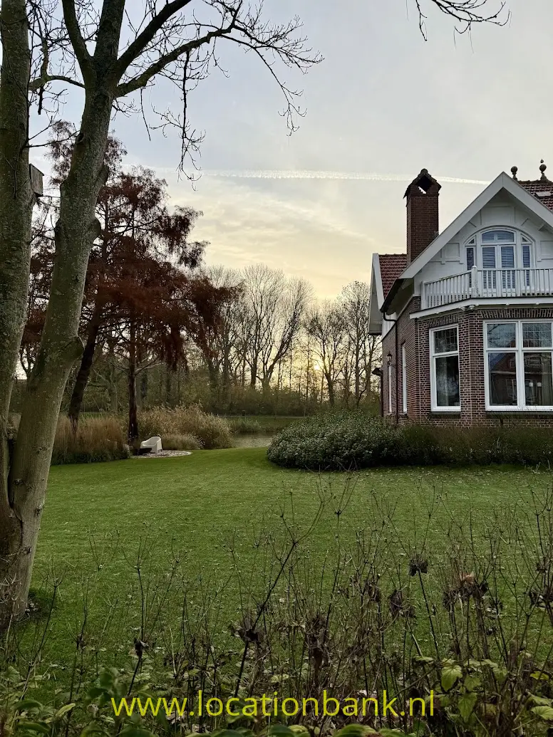 Bakstenen huis met witte balkonbalustrade en puntgevel aan een groen gazon met bomen.