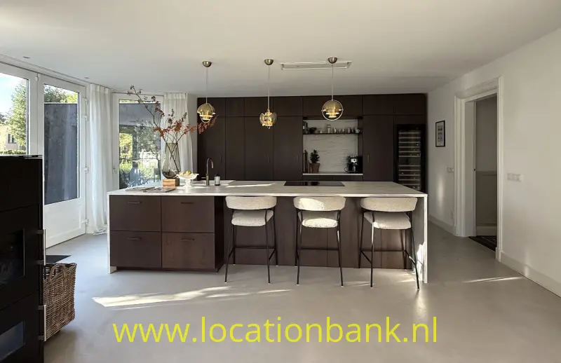 Keuken met een groot kookeiland, drie barkrukken, donkere kasten en drie hanglampen naast hoge ramen.