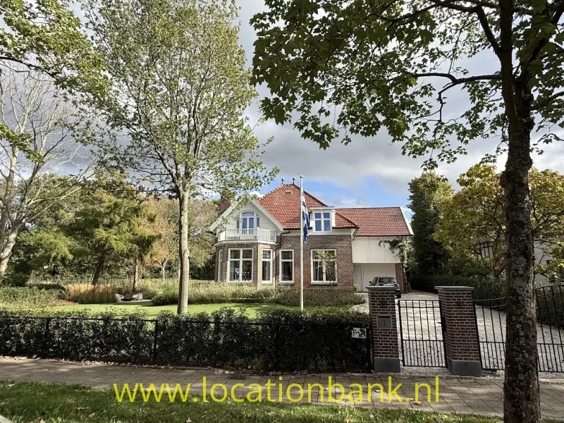 Huis met rode pannendaken, een glazen entree en een afgesloten oprit, omgeven door bomen en een haag.