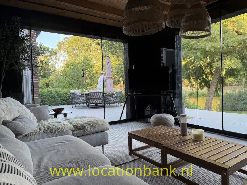 Woonkamer met een grote hoekbank, houten salontafel en kamerhoge ramen met zicht op een tuin en terras.