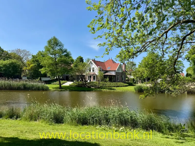 Een huis met rode daken ligt achter een vijver, omgeven door bomen en riet.