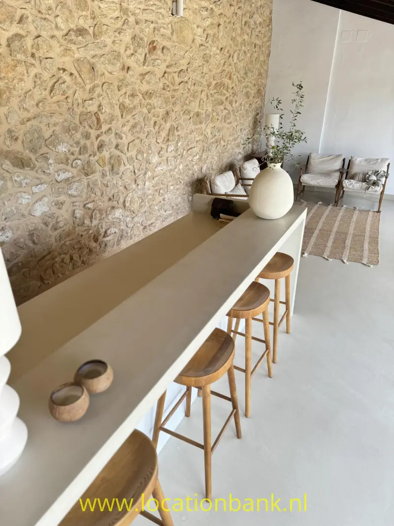 Lange beige bar met houten krukken, een grote vaas en een muur van natuursteen.