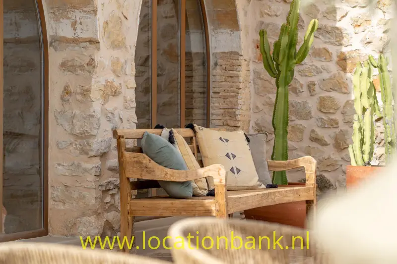 Houten fauteuil met kussens staat tegen een stenen muur, naast hoge cactussen.