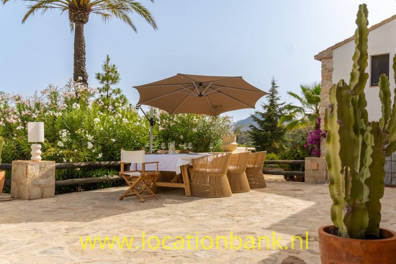 Stenen terras met een eettafel en stoelen onder een grote bruine parasol, omgeven door palmen, bloemen en cactussen.