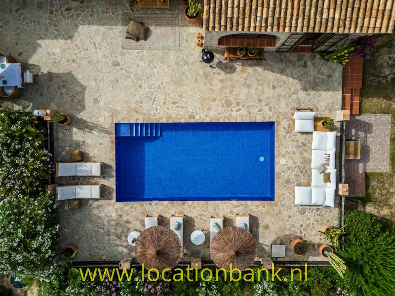 Drone Shot van een Rechthoekig blauw zwembad in een met natuursteen geplaveide binnenplaats, omringd door ligstoelen, parasols en witte zitmeubels.
