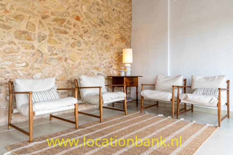 Vier houten fauteuils met witte kussens staan rond een gestreept vloerkleed naast een natuurstenen wand.