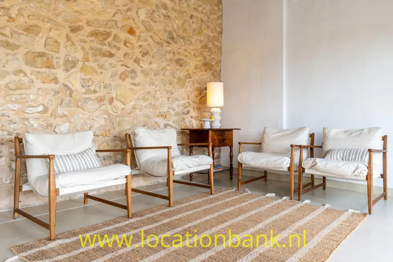 Vier houten fauteuils met witte kussens staan rond een gestreept vloerkleed naast een natuurstenen wand.