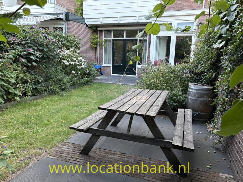 Een houten picknicktafel staat op een betegeld terras in een groene binnentuin naast een bakstenen gebouw.