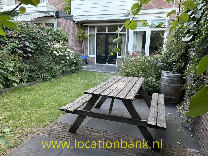 Een houten picknicktafel staat op een betegeld terras in een groene binnentuin naast een bakstenen gebouw.