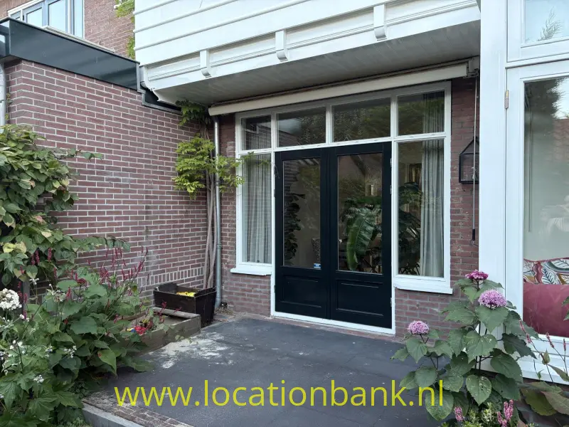 Zwarte dubbele deur in een bakstenen gevel, omgeven door witte raamkozijnen en bloeiende hortensia’s.