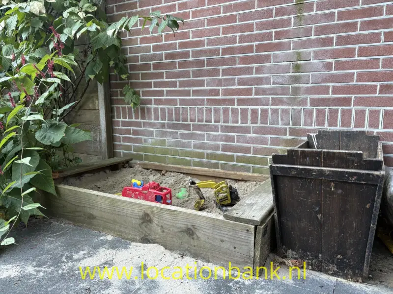 Een zandbak en kleurrijk speelgoed staat tegen een bakstenen muur naast een donkere houten bak.