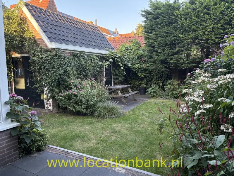 Tuin met een houten picknicktafel, betegeld pad, bloeiende planten en een pannendak op de achtergrond.