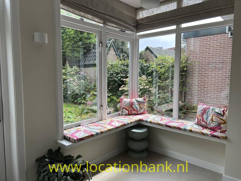 Een hoekraam met een gevoerde vensterbank en kussens met patronen kijkt uit op een groene tuin.