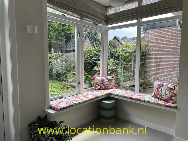 Een hoekraam met een gevoerde vensterbank en kussens met patronen kijkt uit op een groene tuin.