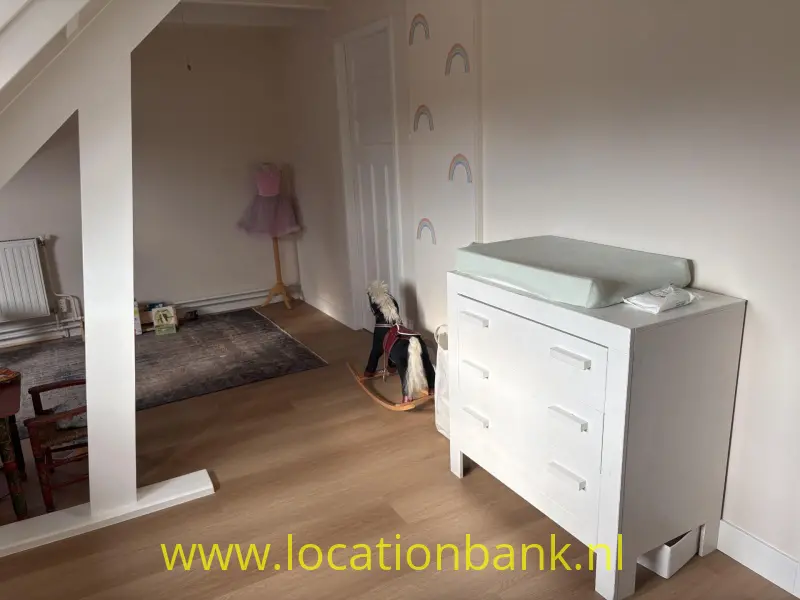 Witte commode met lichtgroen aankleedkussen, regenboogstickers op de muur en een houten hobbelpaard.