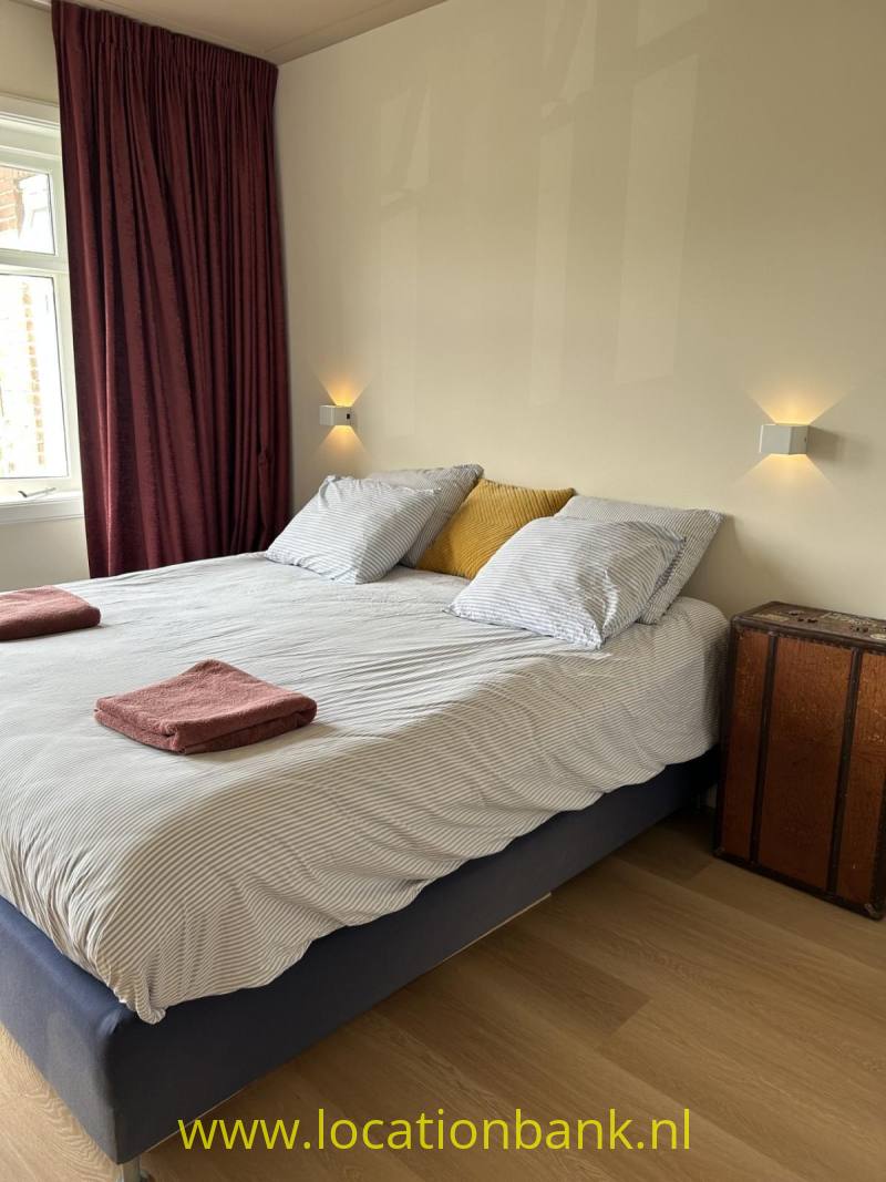 Een tweepersoonsbed met gestreept beddengoed, meerdere kussens en opgevouwen handdoeken staat naast een houten kastje.