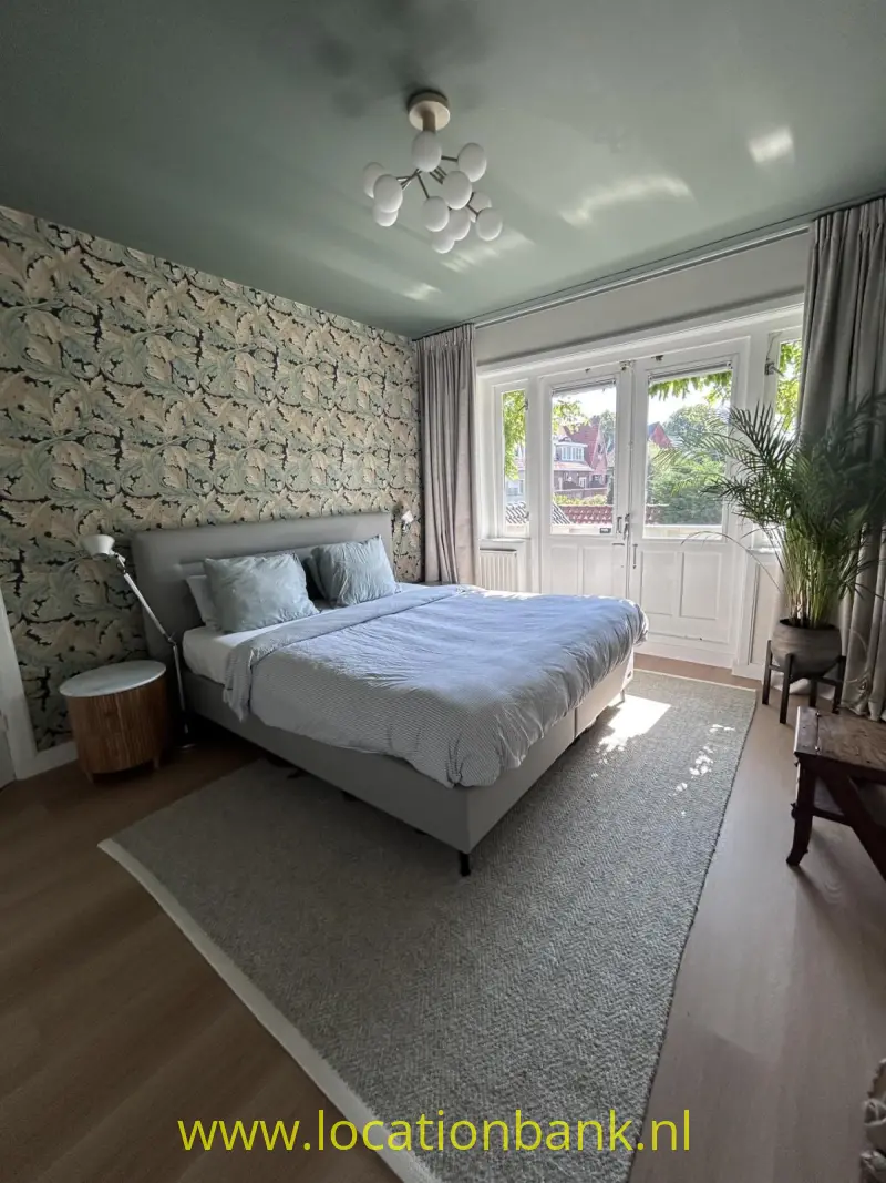 Slaapkamer met een opgemaakt bed, botanisch behang, hoge ramen, gordijnen en een grote kamerplant.