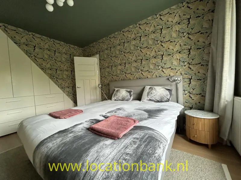 Slaapkamer met een tweepersoonsbed, botanisch behang, witte kastdeuren en een rond houten nachtkastje.