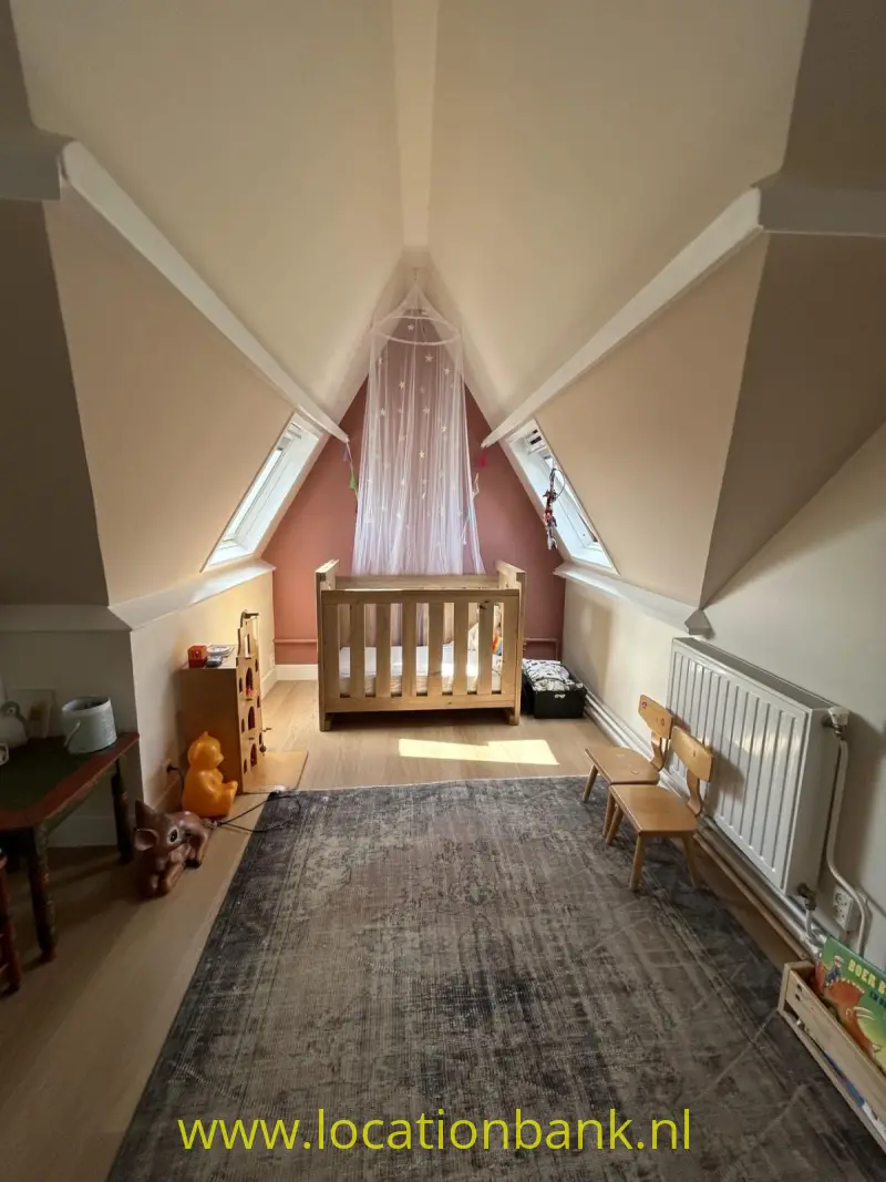 Houten kinderbedje onder een puntig raam in een zolderkamer, met een groot vloerkleed en speelgoed.
