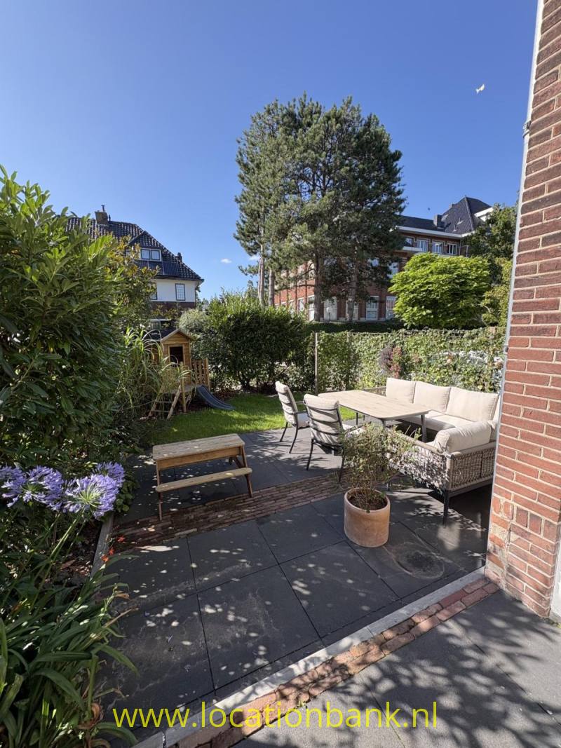 Gemeubileerd terras met een zitbank, tafel, plantenbakken en bloeiende planten in een groene tuin.