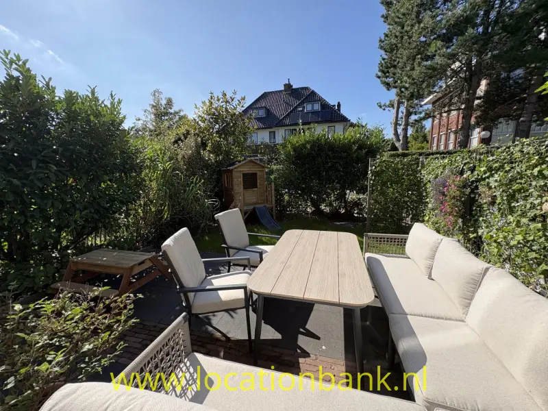 Terras met een lange buitentafel, stoelen en een hoekbank, omgeven door groene hagen en bomen.