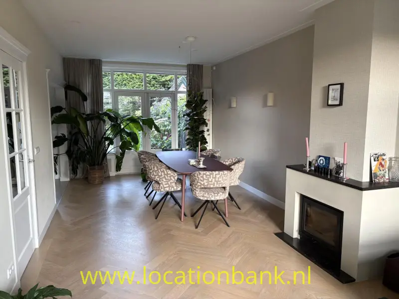 Eetkamer met een ovale tafel, meerdere stoelen, grote ramen, kamerplanten en een haard.