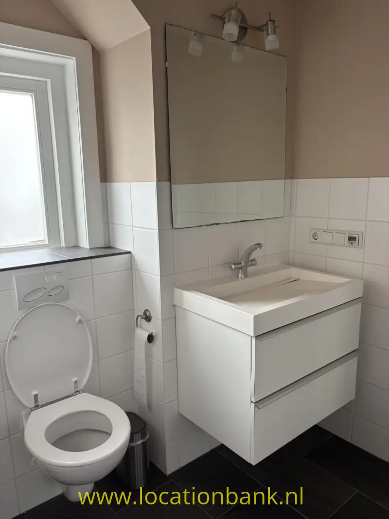 Badkamer met een wit zwevend wastafelmeubel, spiegel, toilet en raam naast betegelde wanden.