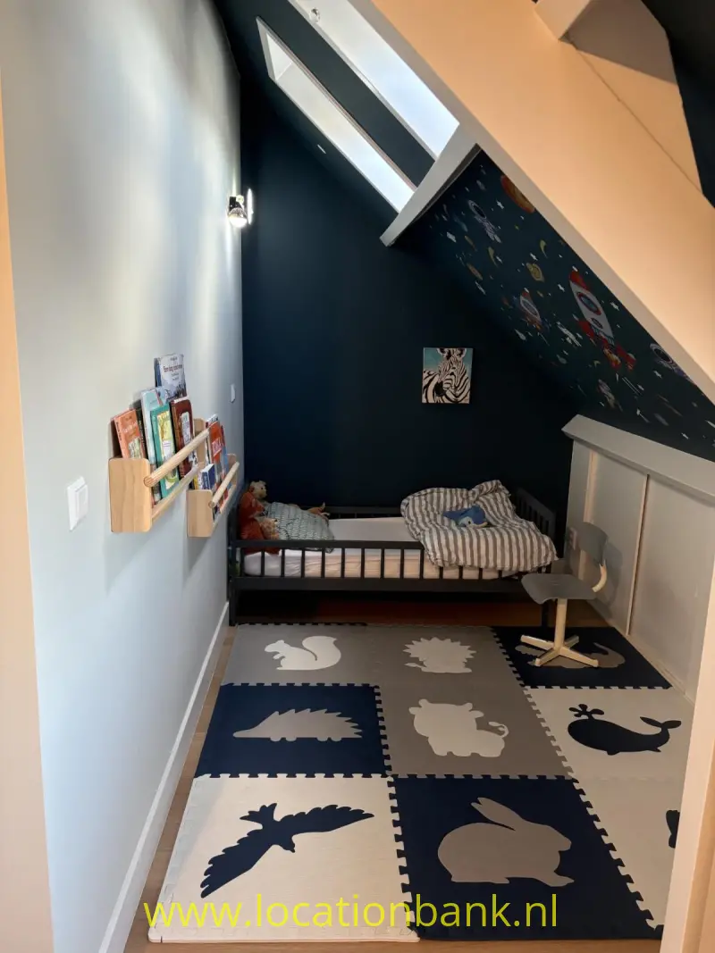 Kinderkamer met een laag houten bed, een dierenpuzzelkleed, een dakraam in het schuine dak en boeken aan de wand.