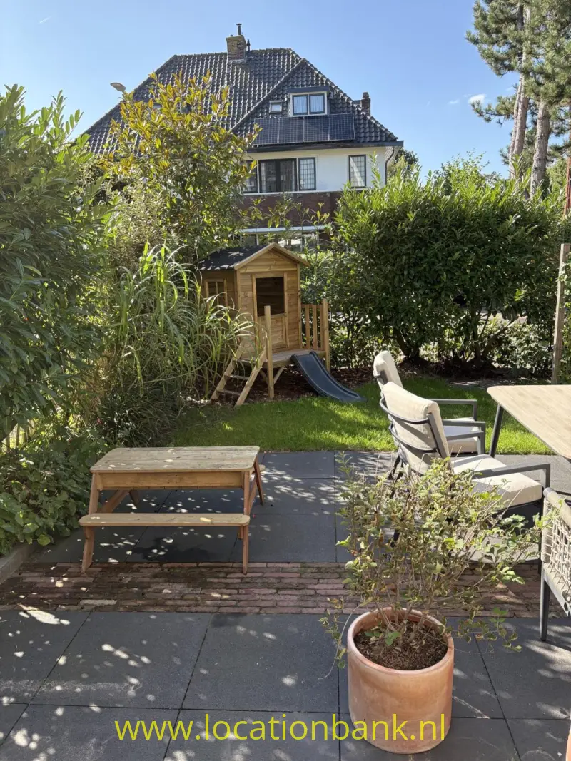 Een houten speelhuisje met glijbaan staat in een groene tuin, met een huis op de achtergrond.