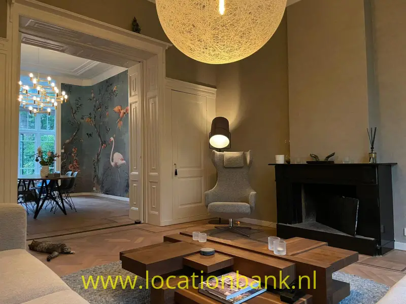 Woonkamer met een zwarte open haard, houten salontafel, fauteuil en aangrenzende eetkamer met gemusterd behang.