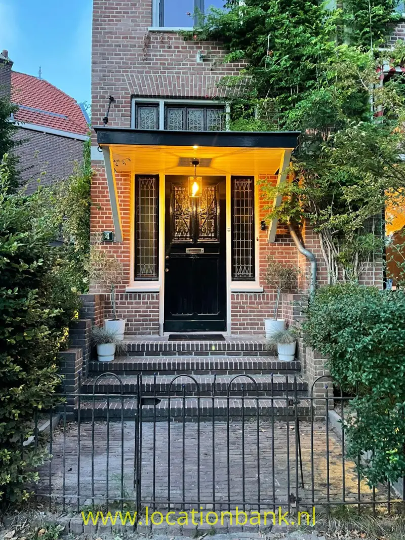 Bakstenen huisentree met een zwarte deur, een gele luifel, traptreden en een metalen hek tussen het groen.