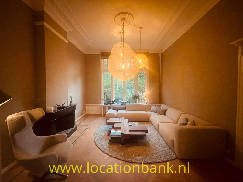 Woonkamer met een beige hoekbank, hanglampen, rond vloerkleed en hoge ramen.