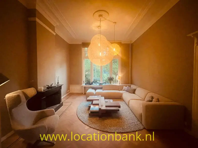 Woonkamer met een beige hoekbank, hanglampen, rond vloerkleed en hoge ramen.