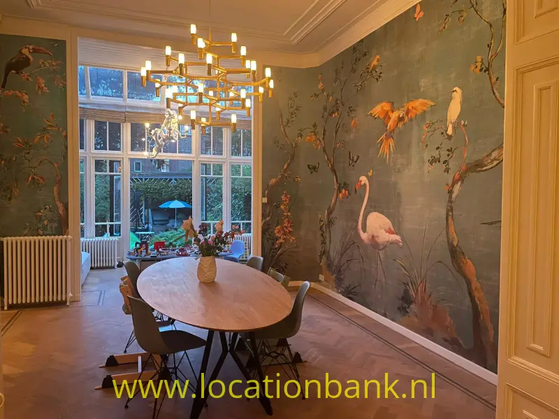 Eetkamer met een ovale houten tafel, een kroonluchter en behang met vogelmotieven.
