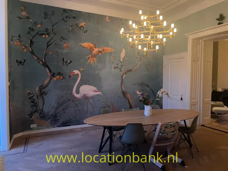 Ruimte met een muurschildering van flamingo’s, vogels en bloeiende takken, een ovale tafel en een kroonluchter.