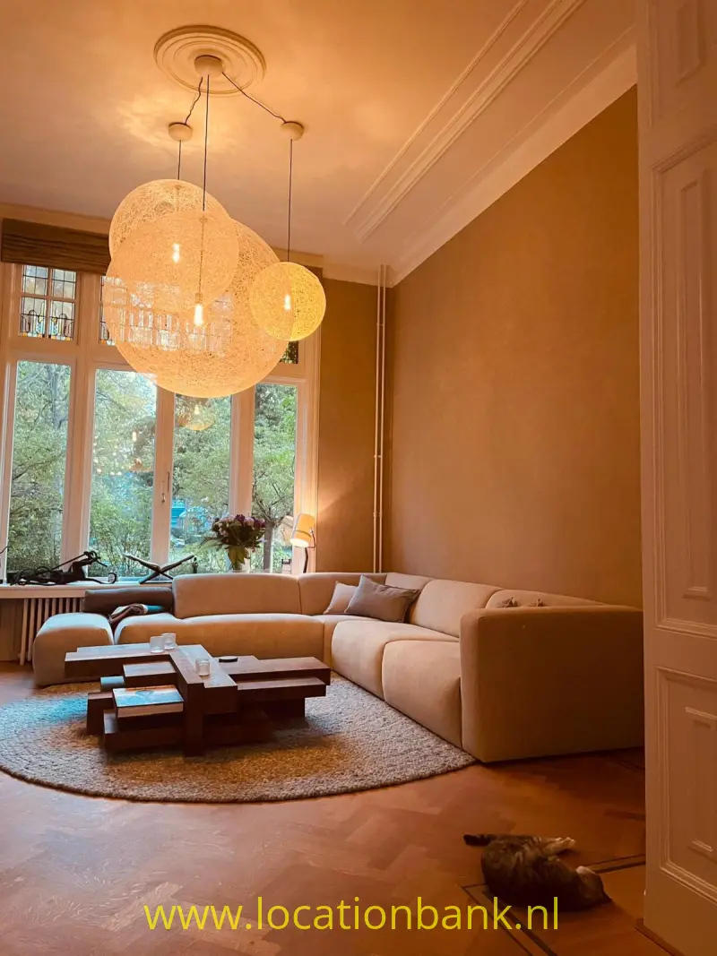 Woonkamer met een gebogen beige hoekbank, drie hangende bolvormige lampen en hoge ramen.
