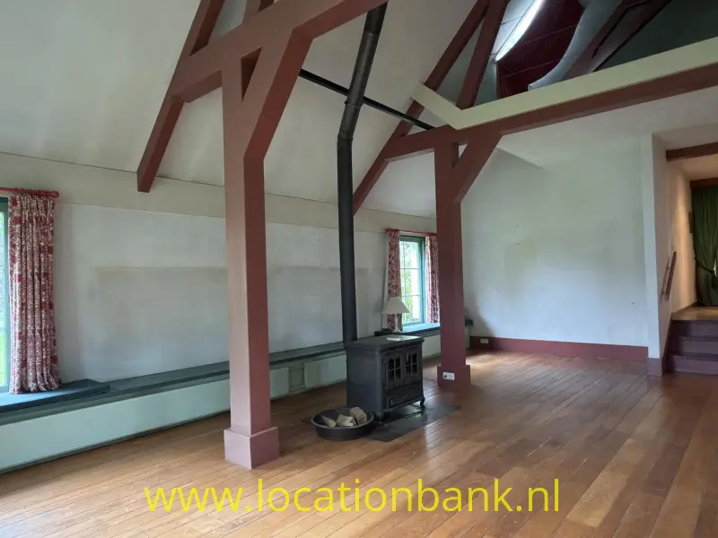 Een zwarte houtkachel staat centraal in een ruime kamer met houten vloer, zichtbare balken en ramen met gordijnen.