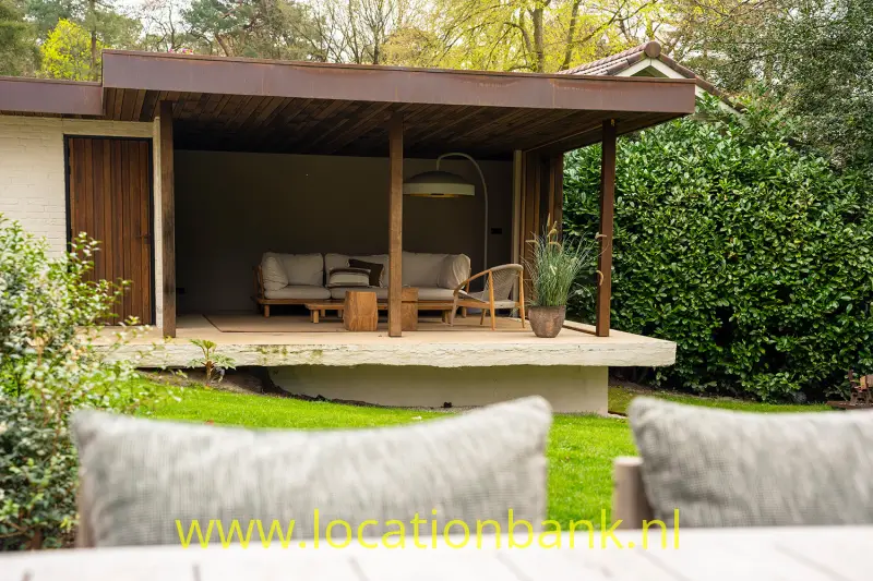 Een overdekt houten terras met zitmeubels staat verhoogd tussen planten, met kussens op de voorgrond.