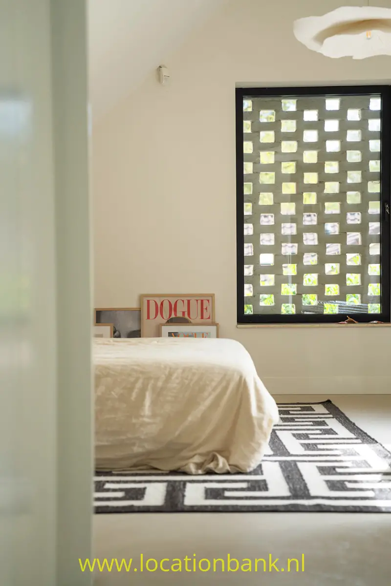 Een bed met wit beddengoed staat op een zwart-witkleed voor een raam met zwarte omlijsting en een raster van vierkanten.