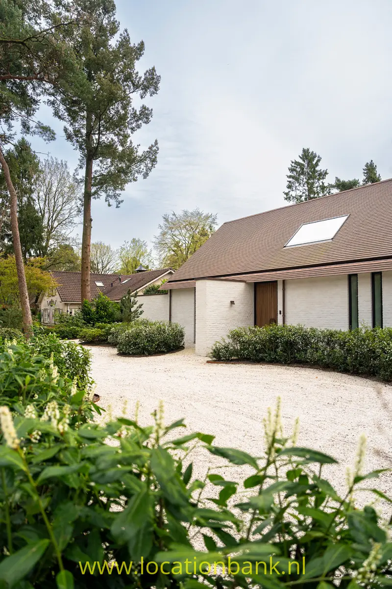 Laag wit gebouw met gebogen pannendak en dakraam aan een met groen omringde grindplaats.