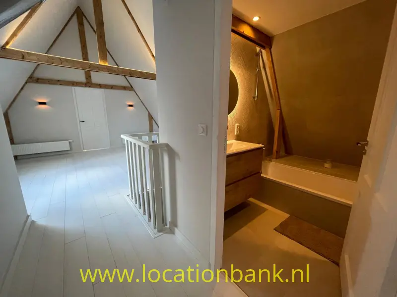 Lichte zolderruimte met zichtbare houten balken naast een badkamer met ligbad.