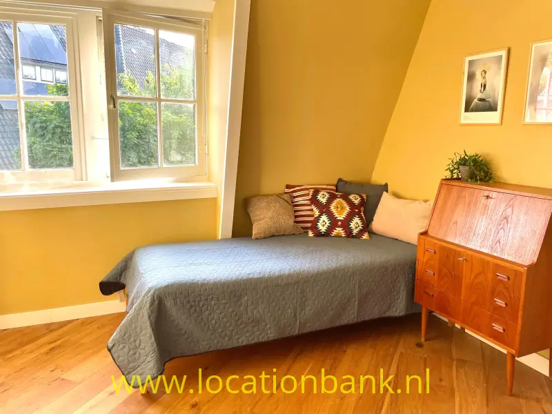 Een bed met een grijze sprei en gedessineerde kussens staat naast een houten ladekast in een gele kamer.