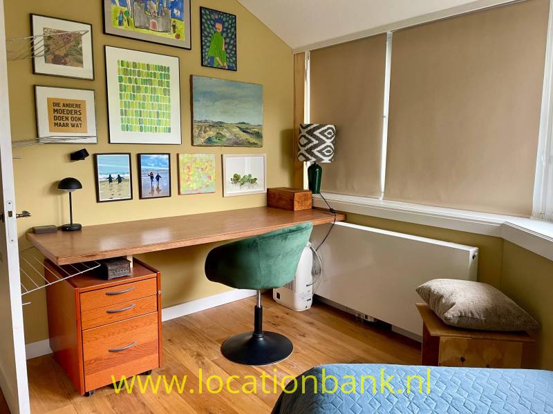 Thuiskantoor met houten bureau, groene bureaustoel, ingelijste kunst en een raam met neergelaten rolgordijnen.
