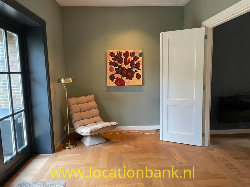 Zithoek met een loungestoel, vloerlamp, ingelijst bloemenschilderij en visgraatvloer naast een open witte deur.