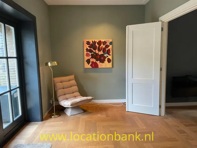 Zithoek met een loungestoel, vloerlamp, ingelijst bloemenschilderij en visgraatvloer naast een open witte deur.