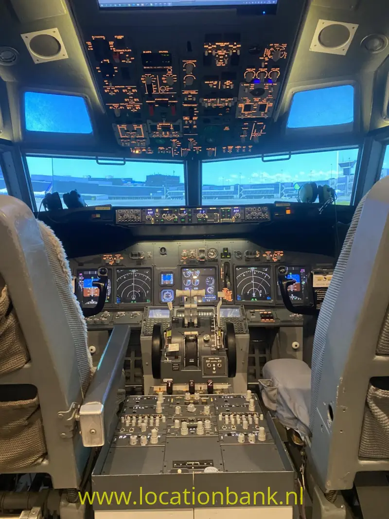 De cockpit toont bedieningspanelen, besturingswielen en een verlicht bovenpaneel, met vliegveld infrastructuur zichtbaar door de ramen.
