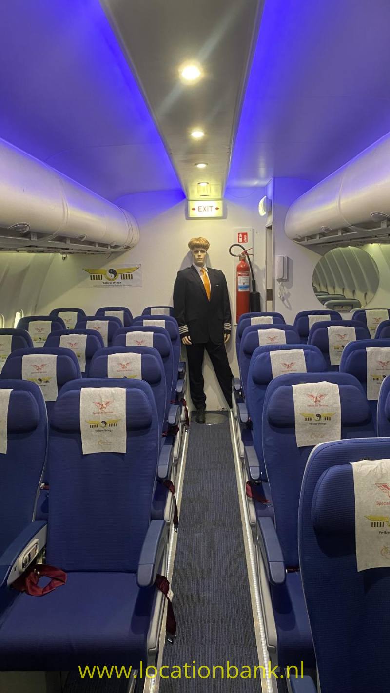 Een stewart staat in het gangpad van een vliegtuigcabine tussen rijen blauwe stoelen en open bagagevakken.