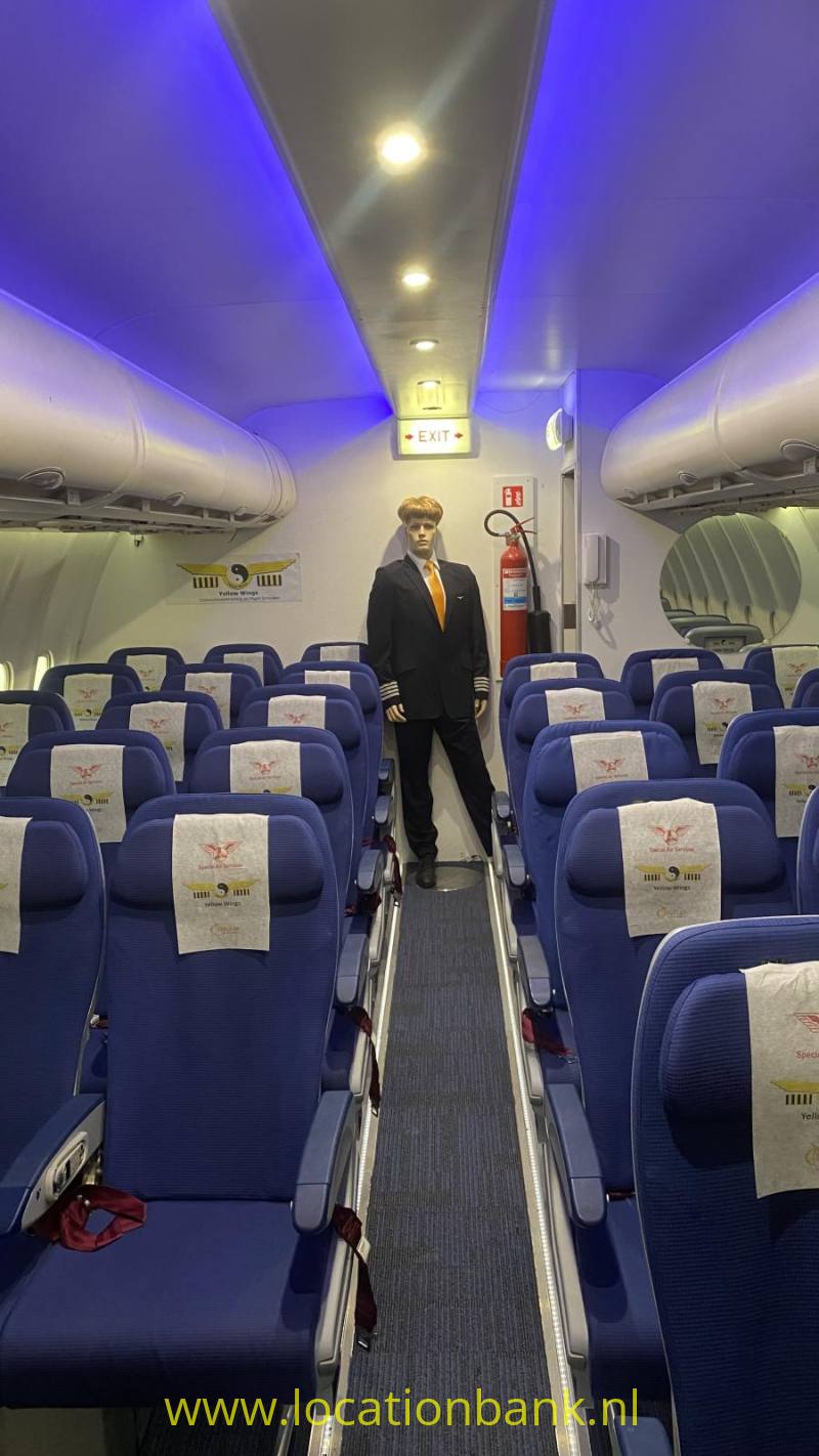 Een man in pak staat in het gangpad van een vliegtuig tussen rijen blauwe stoelen en blauwe verlichting.