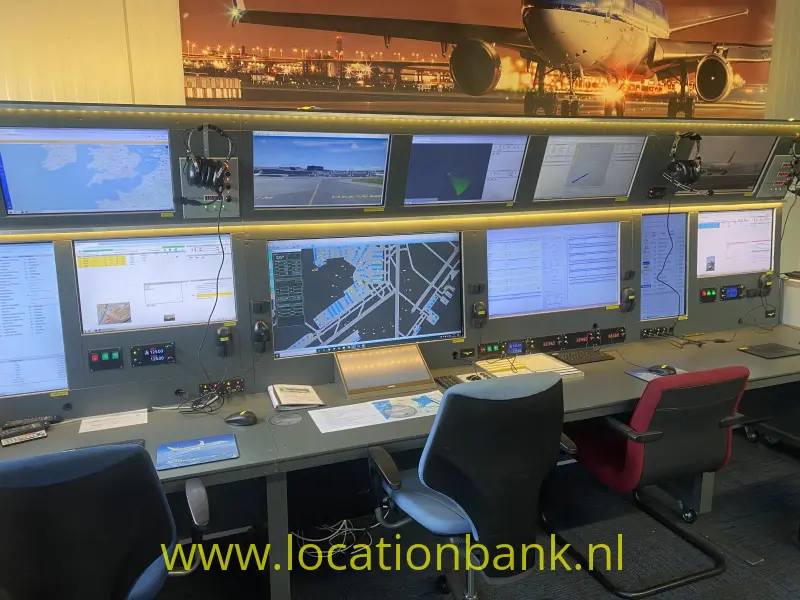 Een luchtverkeers leiding met meerdere computerschermen, bedieningspanelen en bureaustoelen onder een groot vliegtuigbeeld.